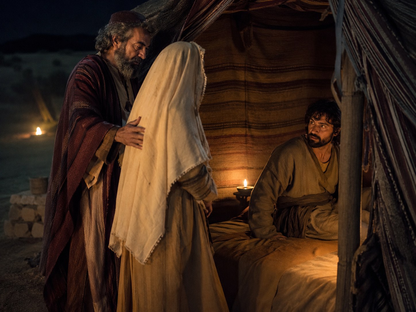Interior escuro de tenda ao anoitecer, uma lamparina de azeite quase apagada sobre uma pedra, e vultos apenas insinuados no fundo.